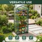 A freestanding multi-tier greenhouse with a transparent green frame stands on a wooden deck in a landscaped garden. The greenhouse holds several potted flowers, vegetables, and gardening tools.